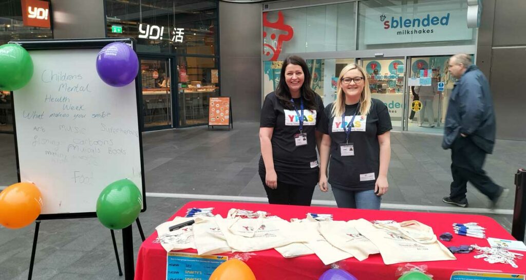 Two women standing at a table showing leaflets, with balloons decorating