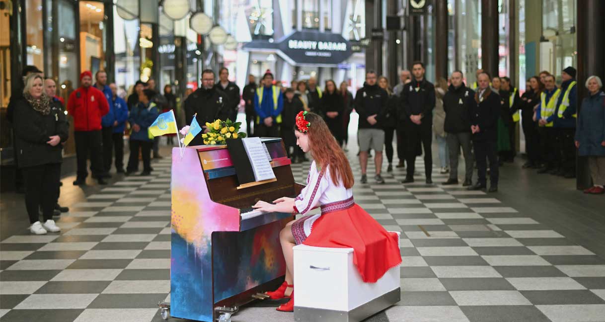 A girl in Ukrainian traditional dress plays piano in the foreground while people assemble