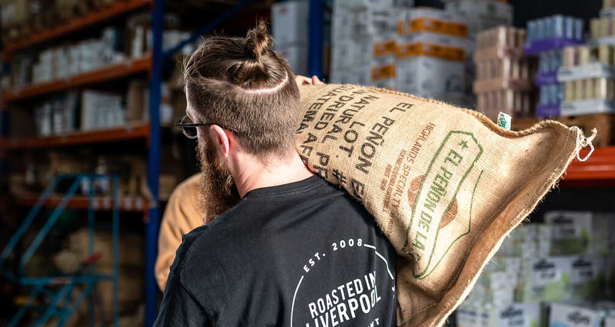 a man from behind carrying a coffee bean sack with "brewed in liverpool" on his tshirt