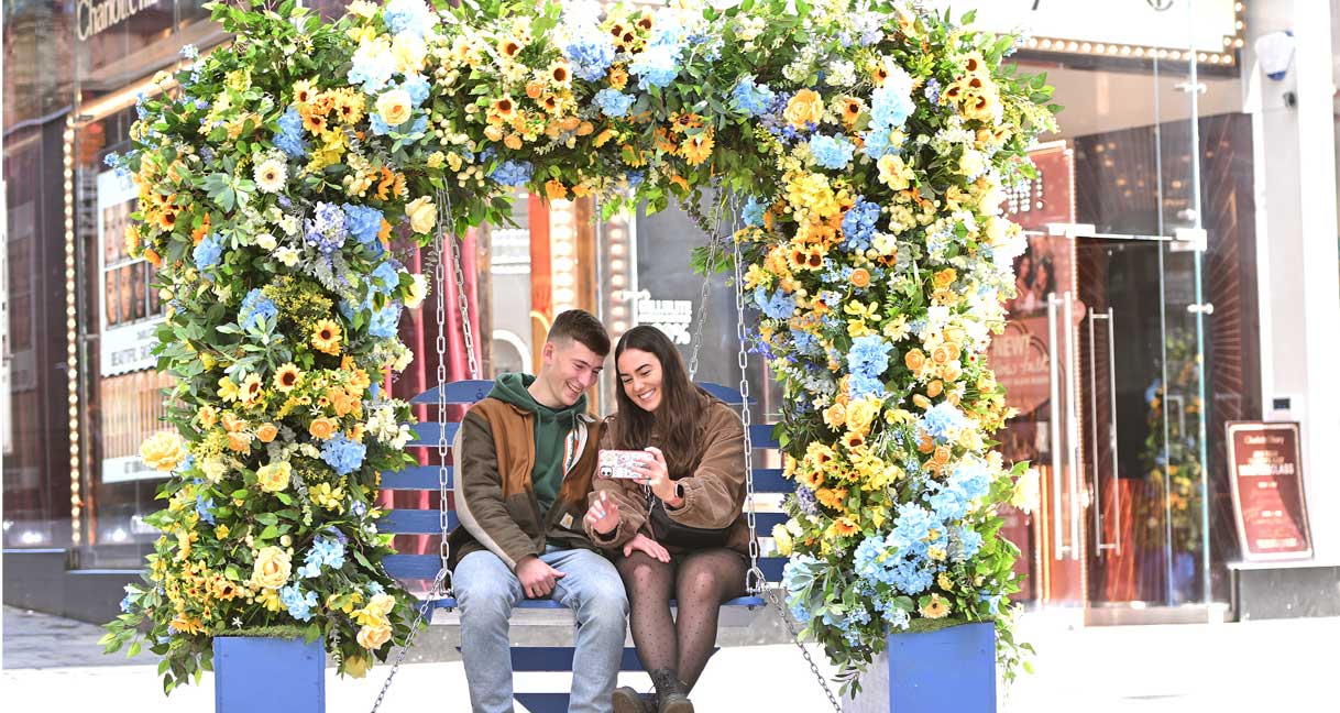 a couple sit in the Liverpool ONE floral installation swing looking at a phone