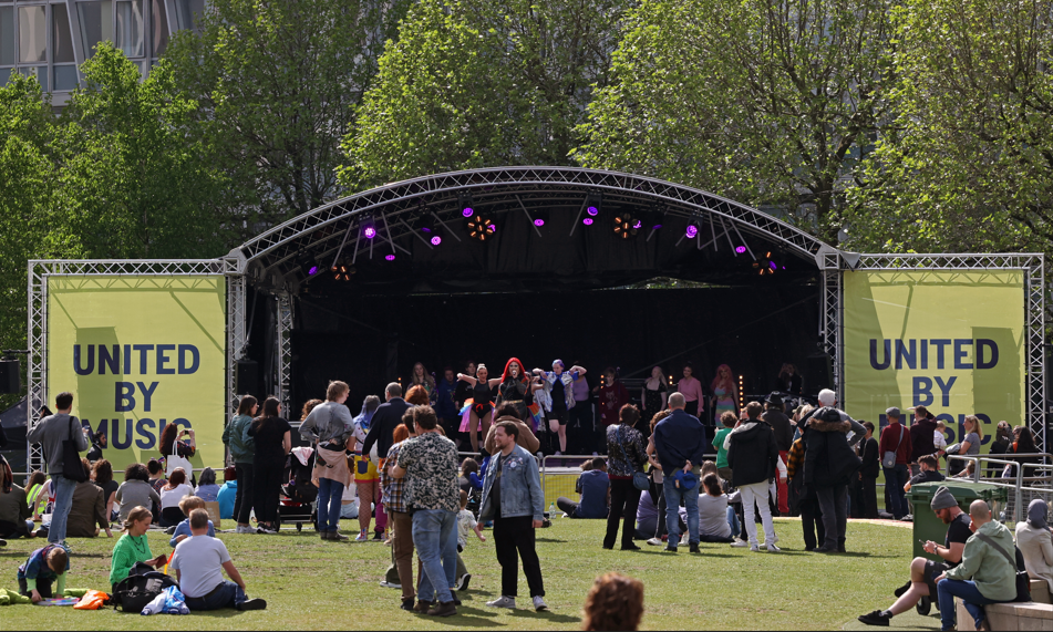 A wide shot of a stage on the park featuring a drag performance and spectators on a sunny day